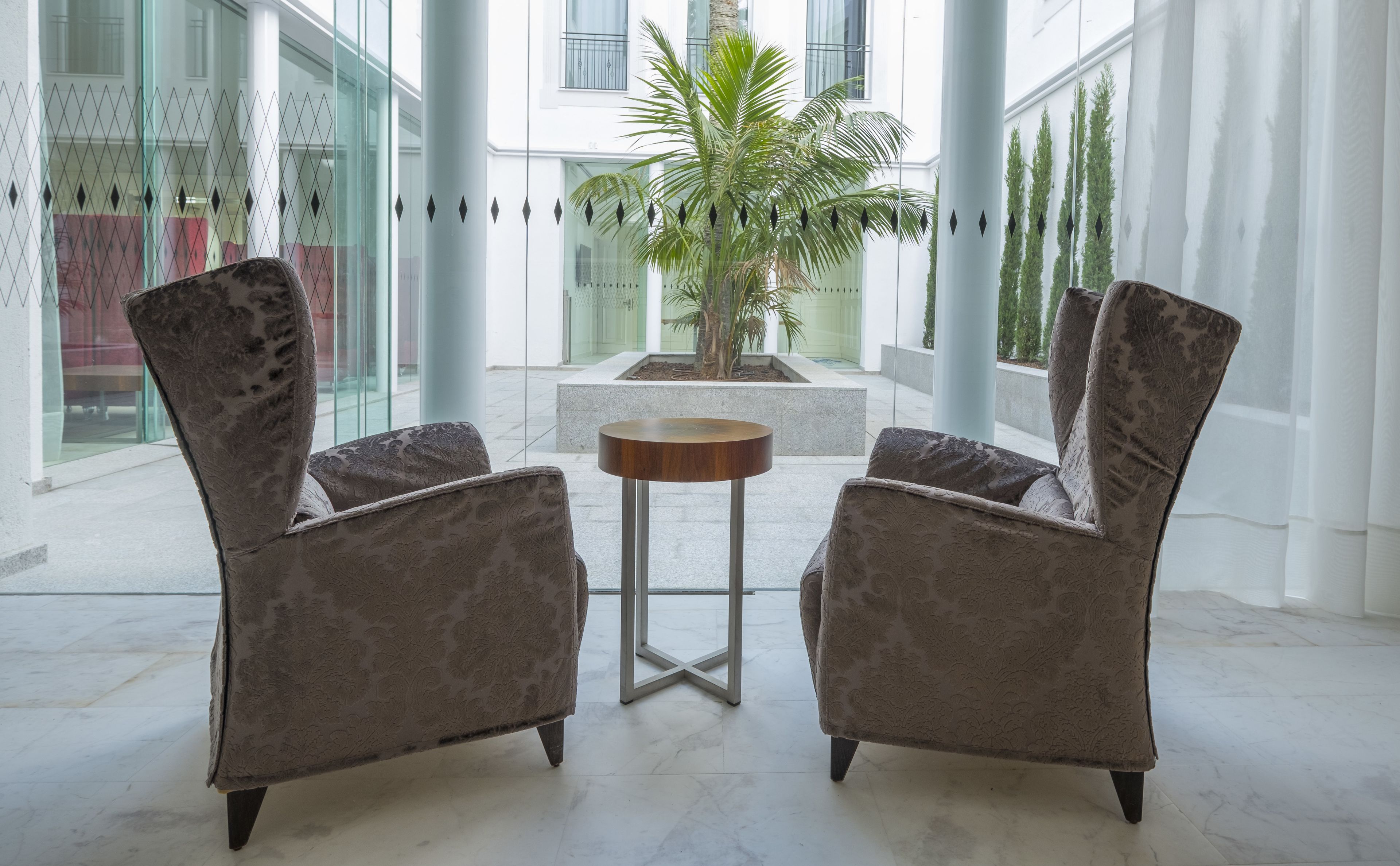 A modern interior with two upholstered chairs facing a small wooden table, a palm tree, and a glass wall with a geometric pattern.