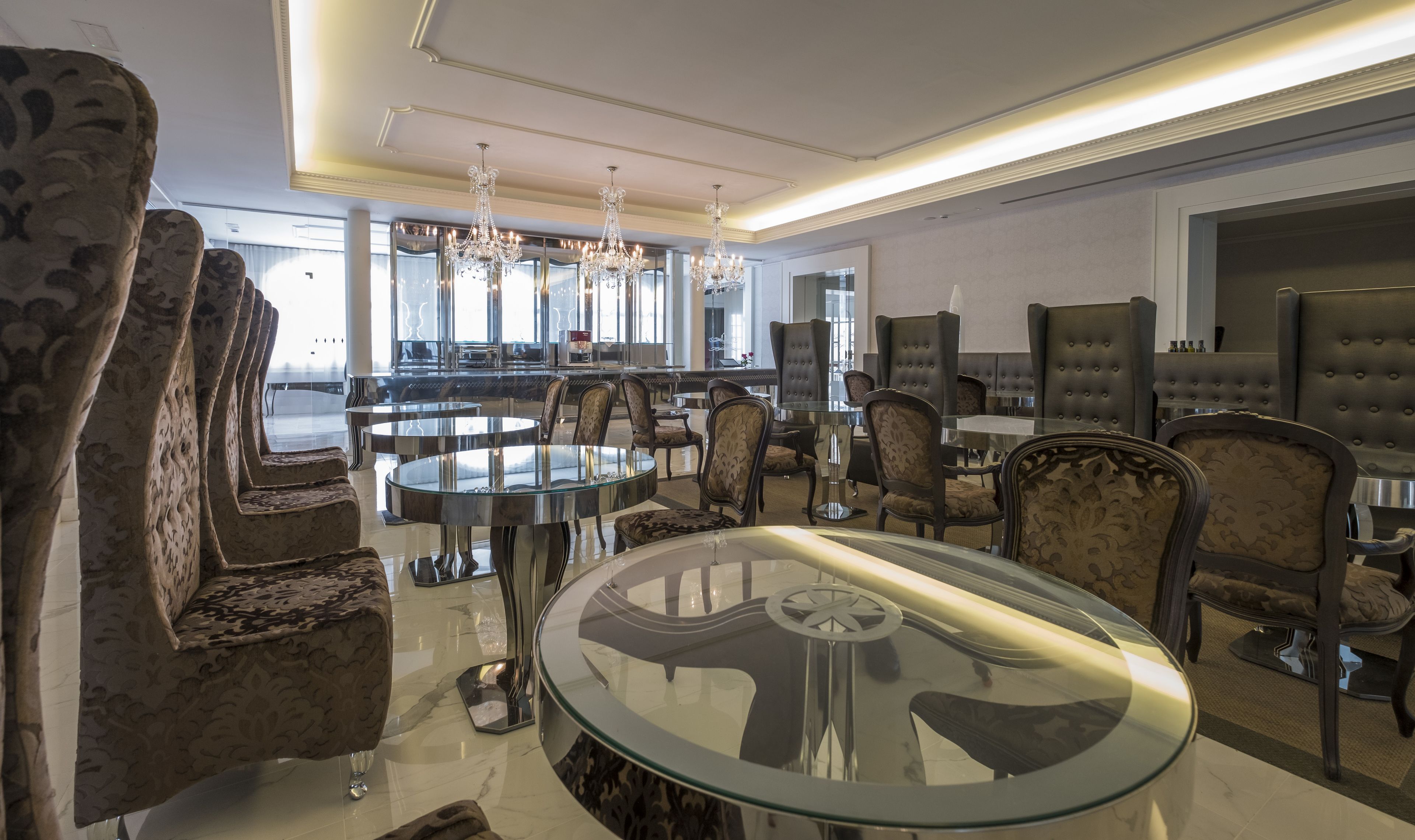 Een moderne eetruimte met ronde glazen tafels en gestoffeerde stoelen, een kroonluchter en een uitzicht op de stad door grote ramen.