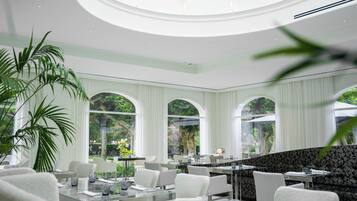 A modern dining area with glass tables, white chairs, and large windows offering views of greenery.