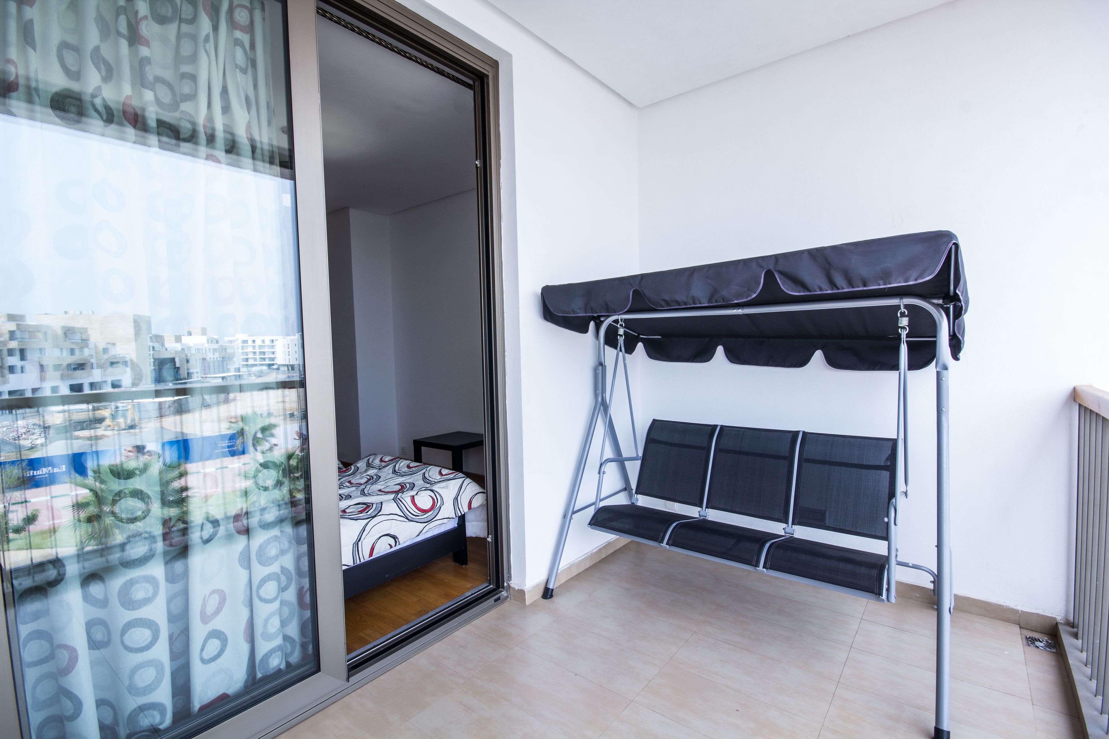 A balcony with a bed, a window with patterned curtains, and a view of buildings.