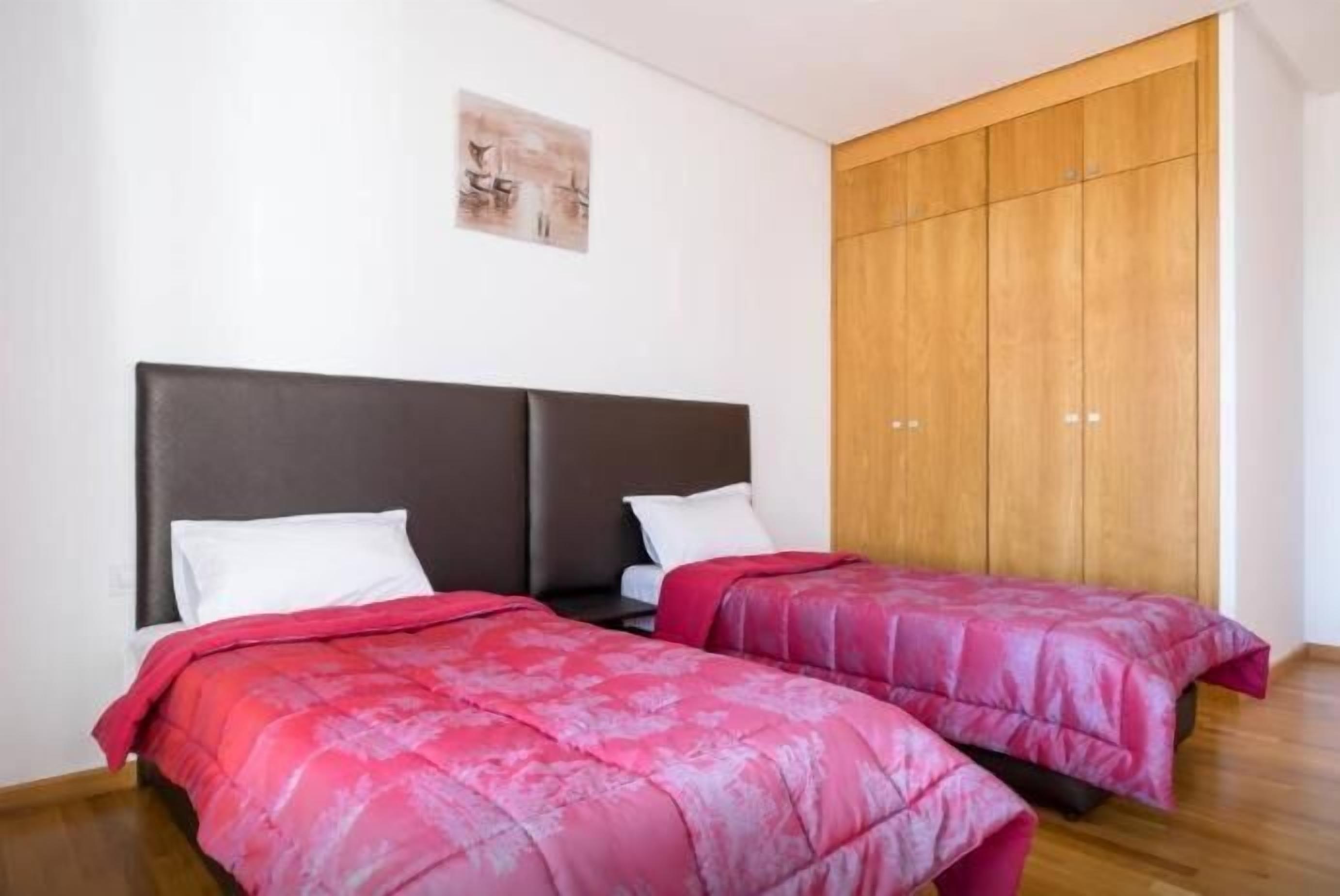 Una habitación con dos camas, cada una con un cabecero oscuro y ropa de cama roja. Se observa un armario de madera y un pequeño cuadro en la pared.