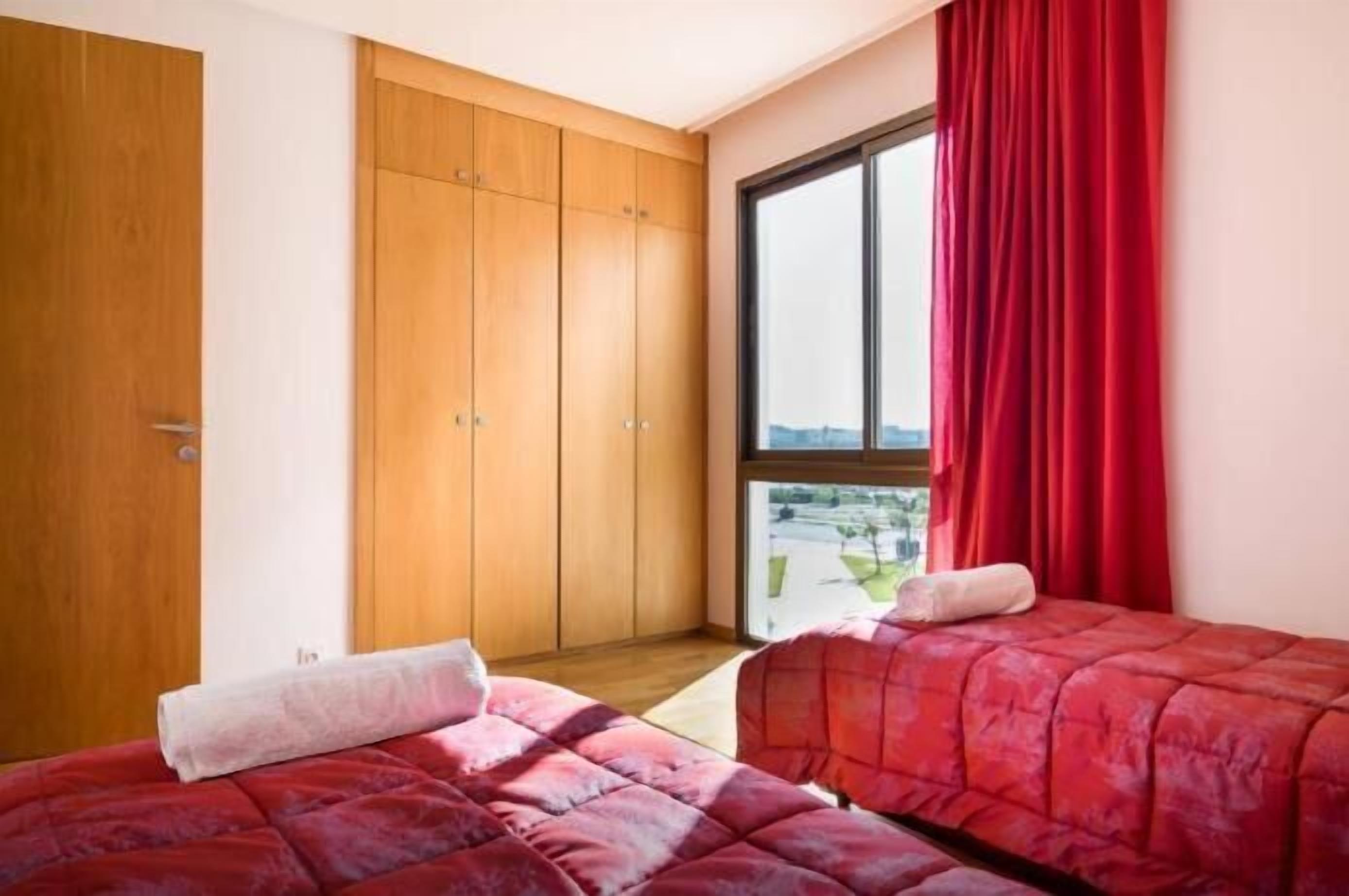 A bedroom with two beds, wooden wardrobe, and a large window with red curtains.