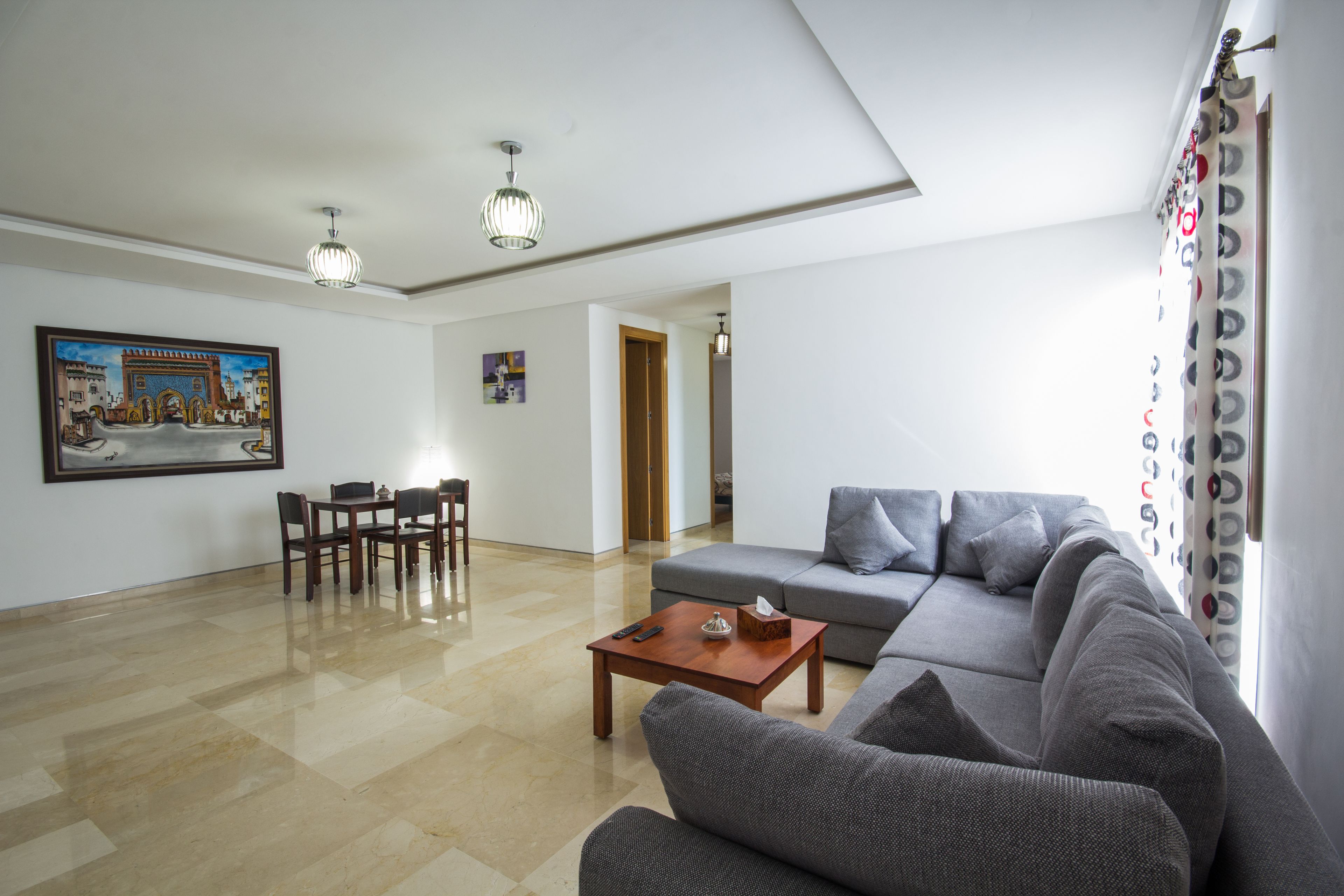 A living room with a grey sofa, a wooden coffee table, a dining table with chairs, and a painting on the wall.