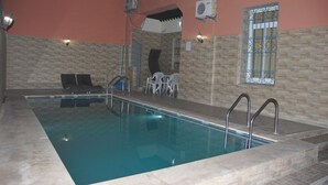 Una piscina interior con pared de azulejos, una ventana con celosías decorativas y un área de estar con sillas.