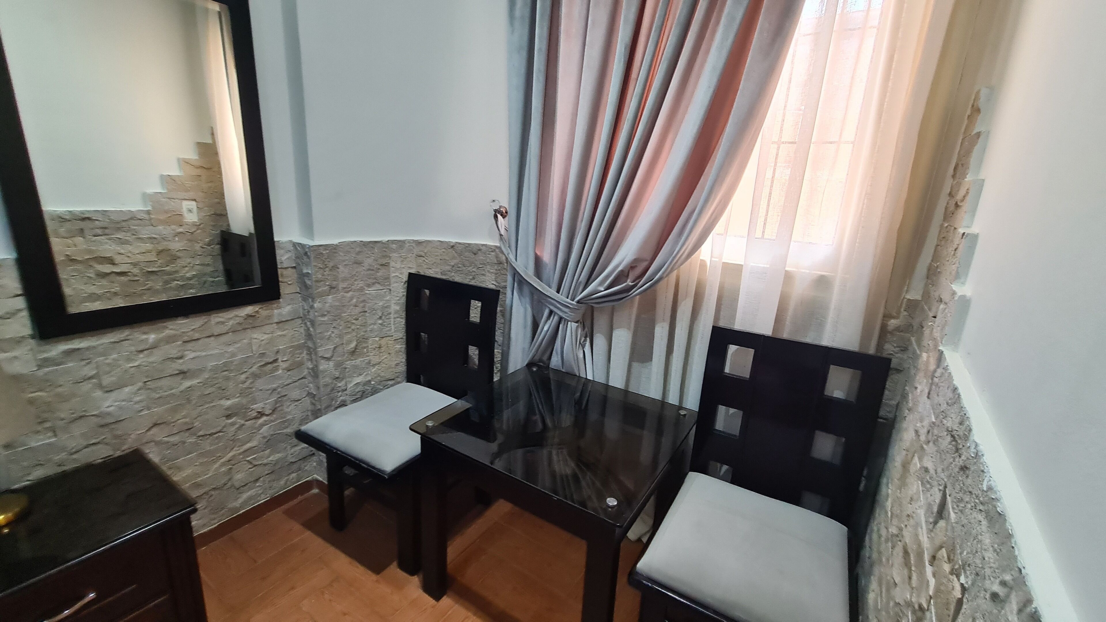 Um quarto pequeno com parede de pedra, um espelho, uma mesa com duas cadeiras e uma janela com cortinas.