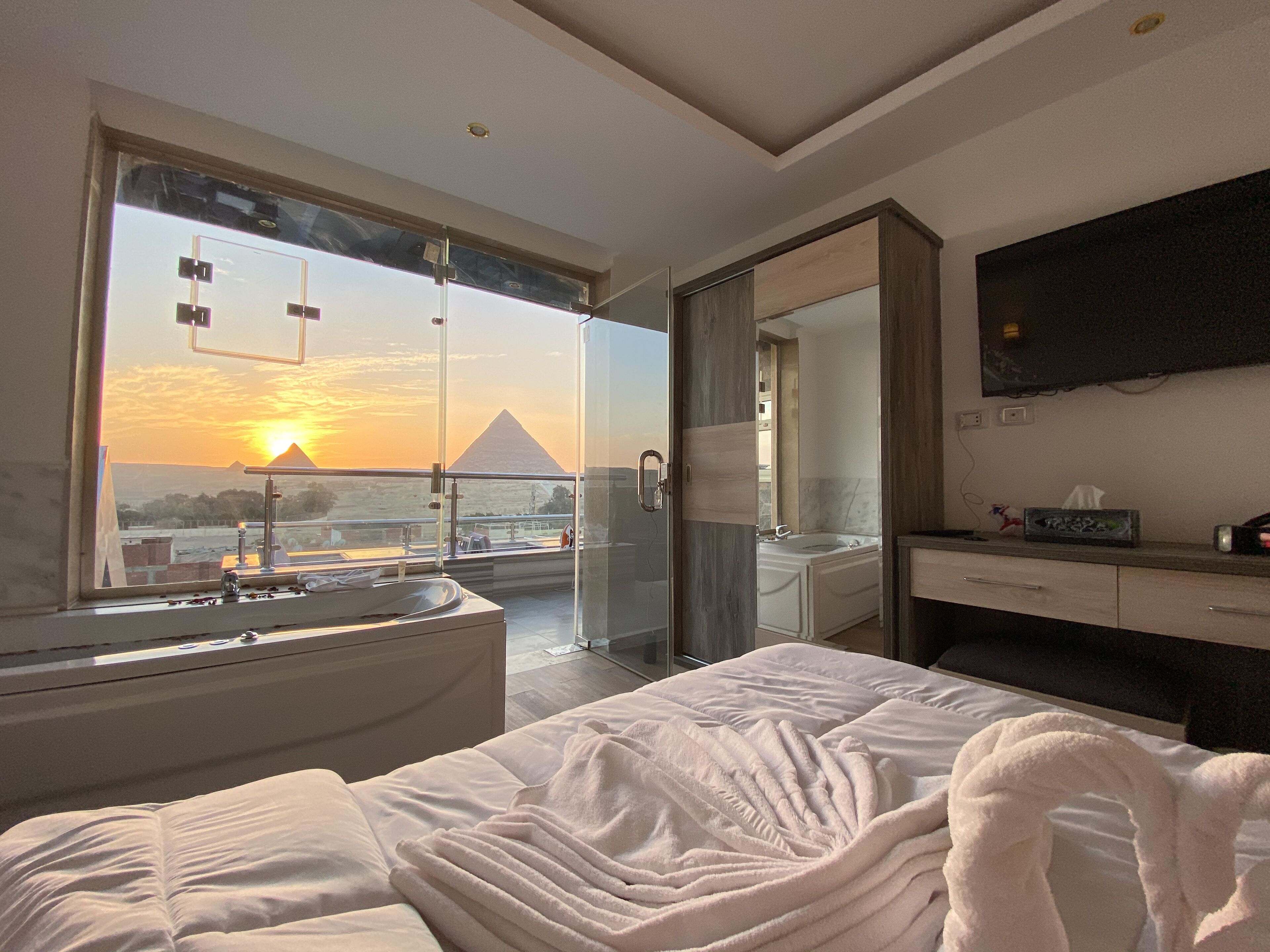 Een moderne hotelkamer met een groot bed, een badkamer met bad en uitzicht op de piramide bij zonsondergang.