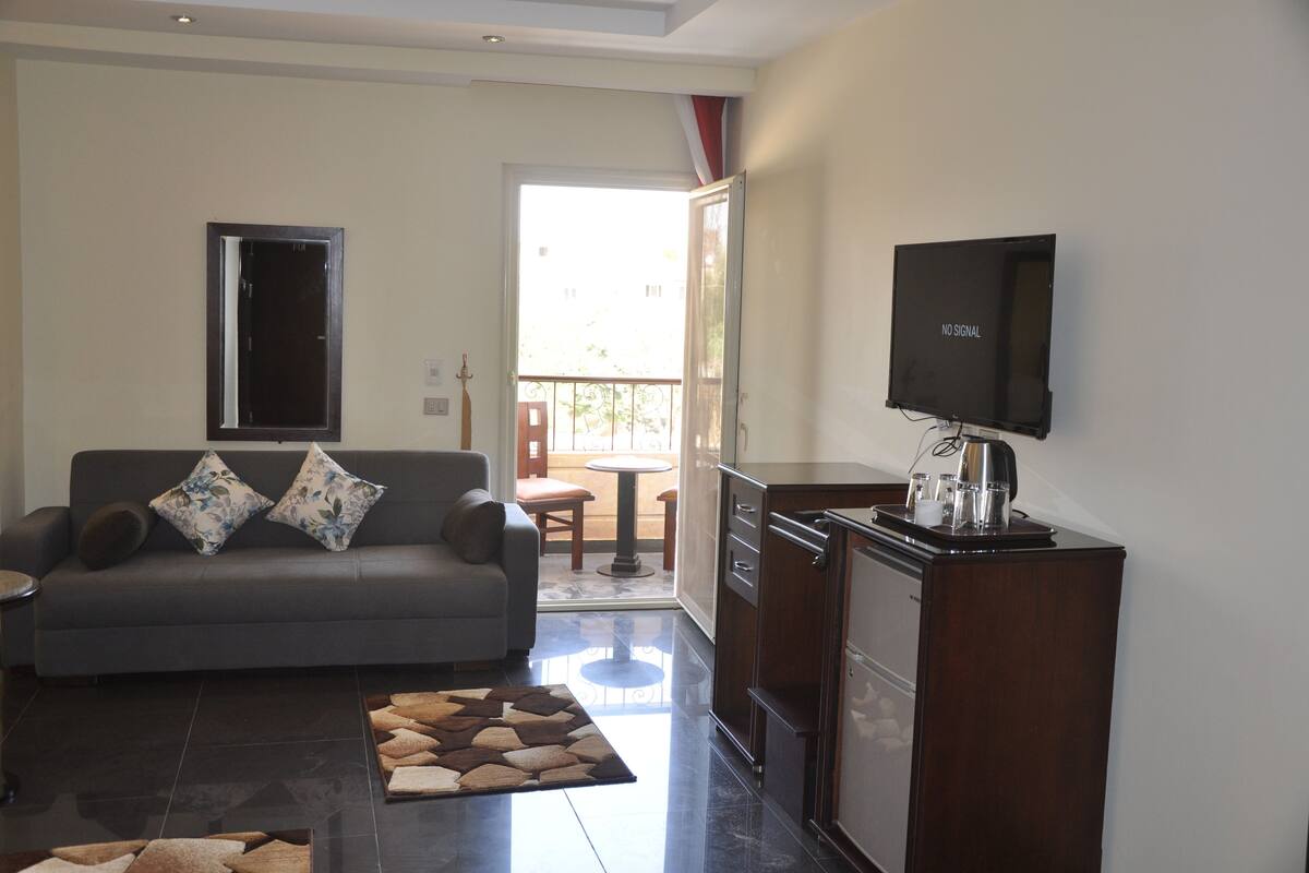 A living room with a sofa, a coffee table, a TV, and a kitchen area with a refrigerator.
