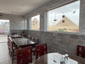 A restaurant with a long table and chairs, large windows with a view of pyramids, and stone walls.