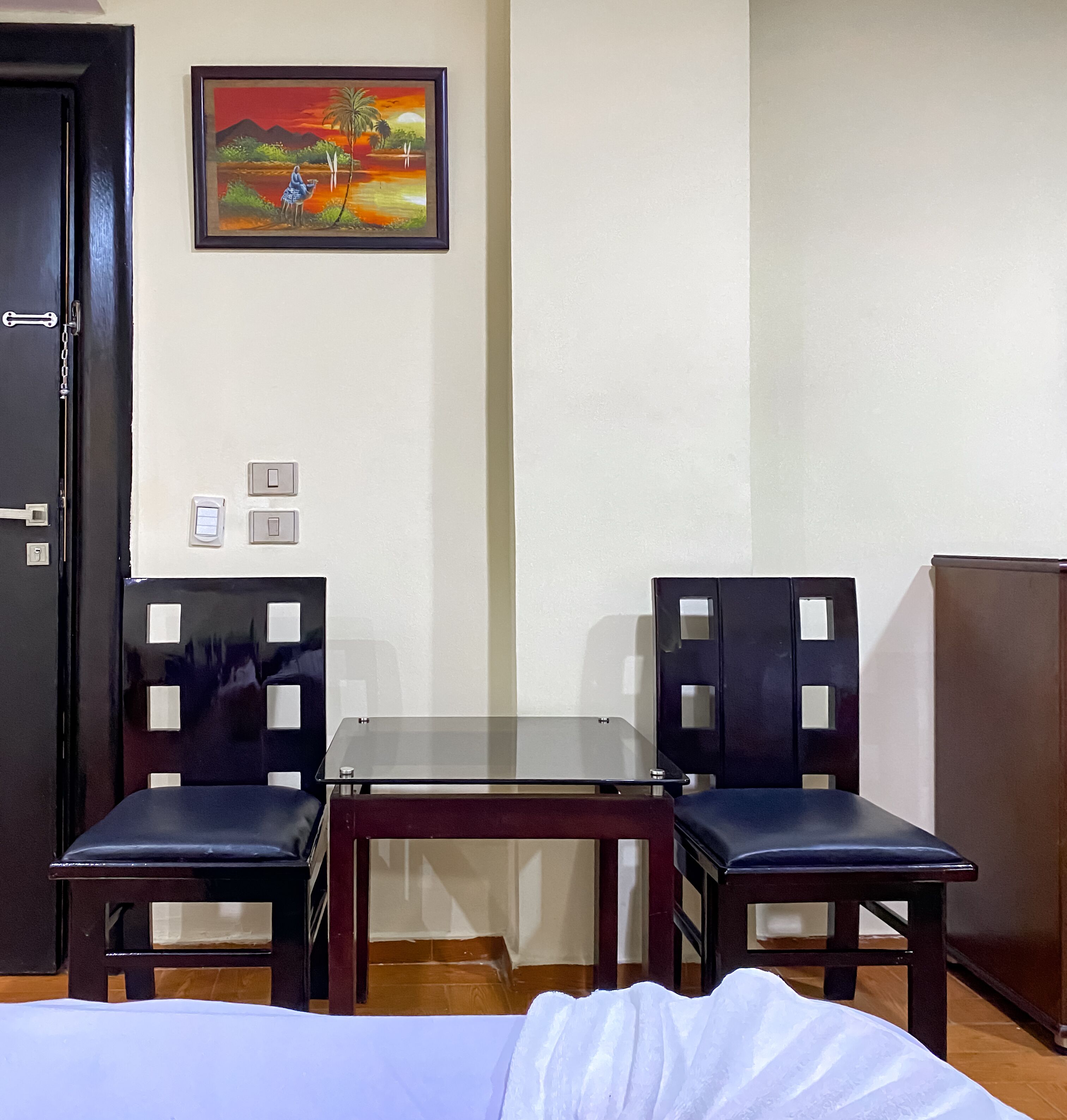 A hotel room with a bed, two chairs, a small table, a painting on the wall, and a door.