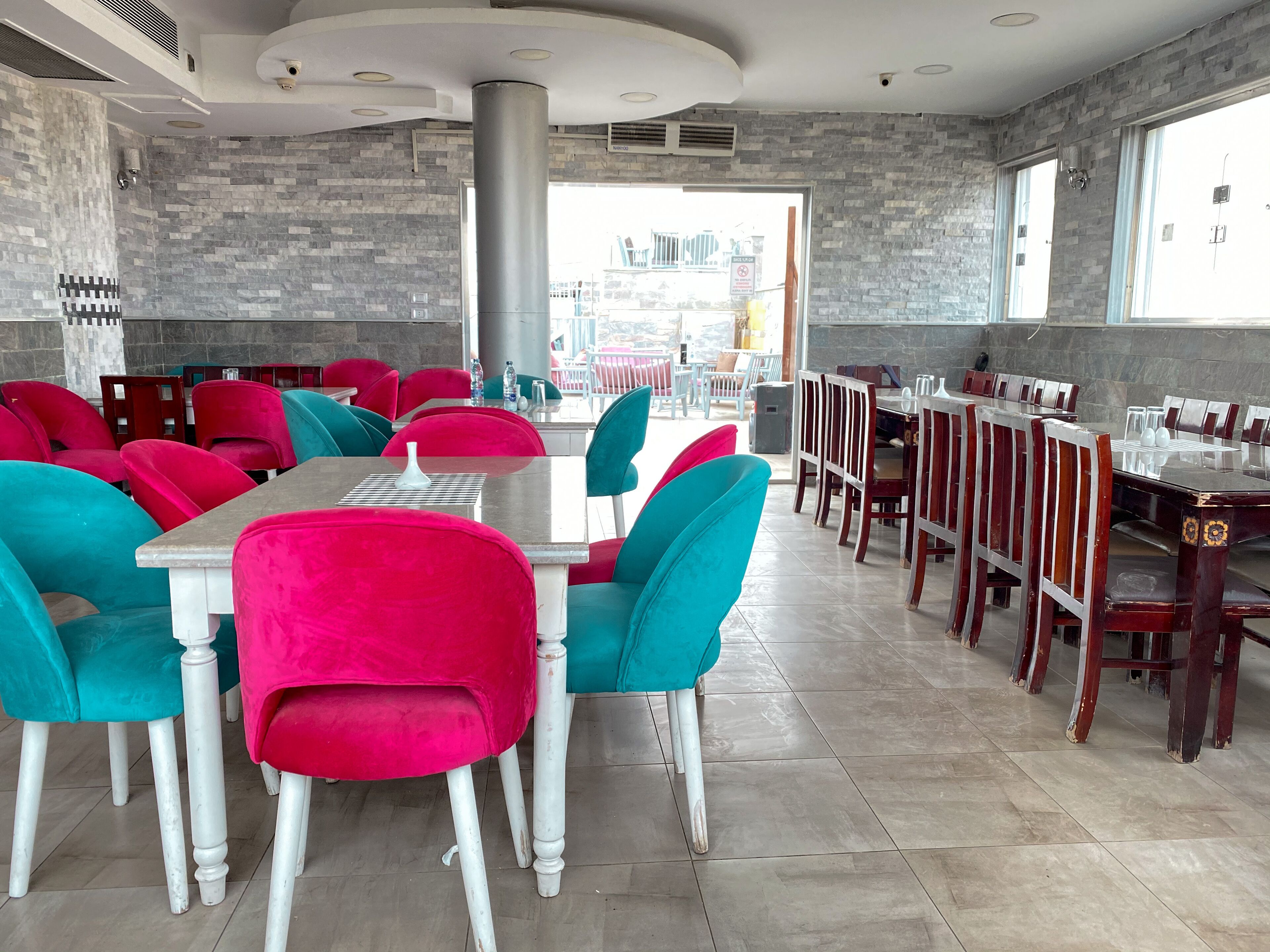 A restaurant with red and teal chairs, white tables, and a stone wall.