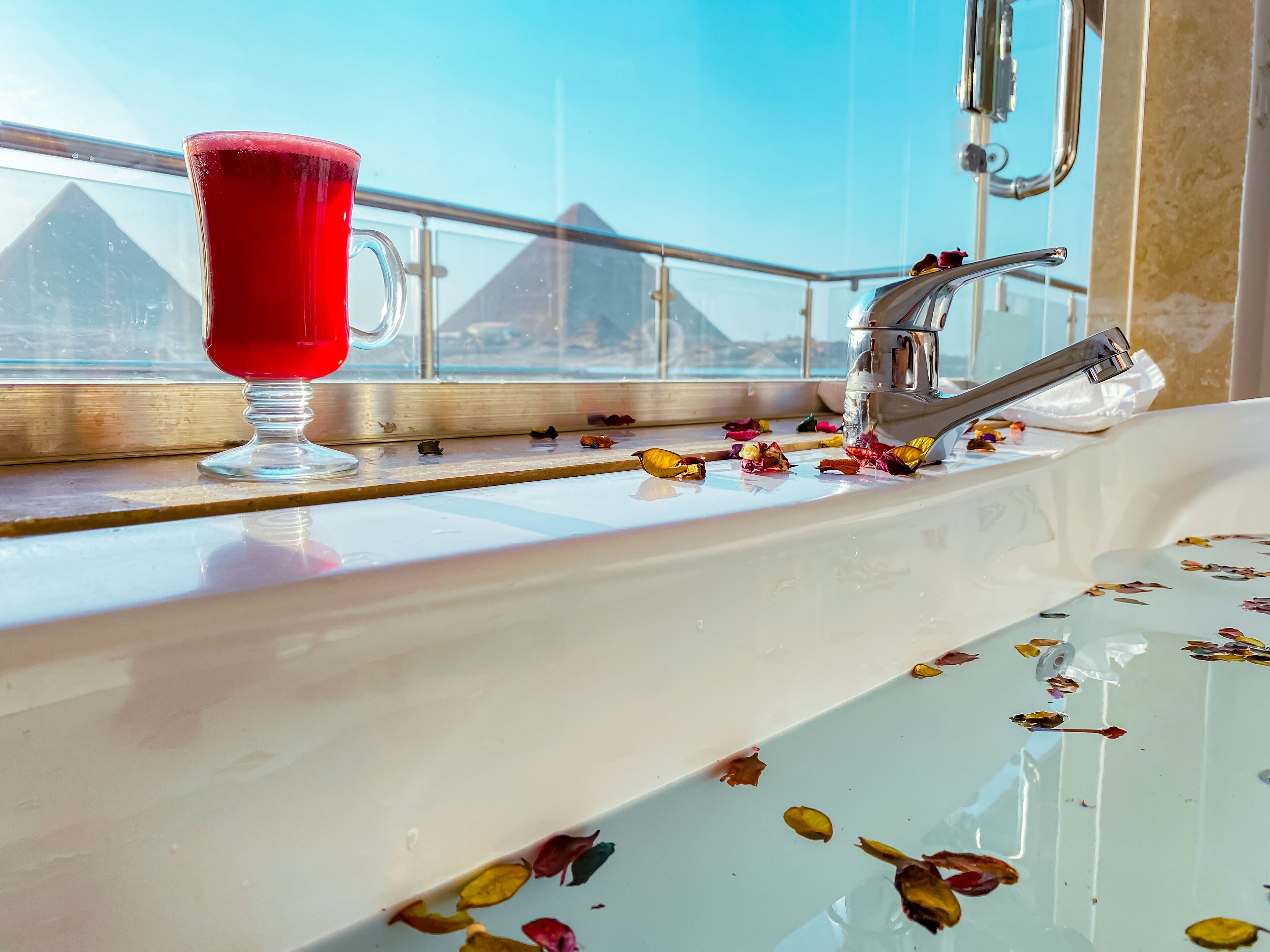 A bathroom with a sink, a glass of red drink, and a window with a view of pyramids.