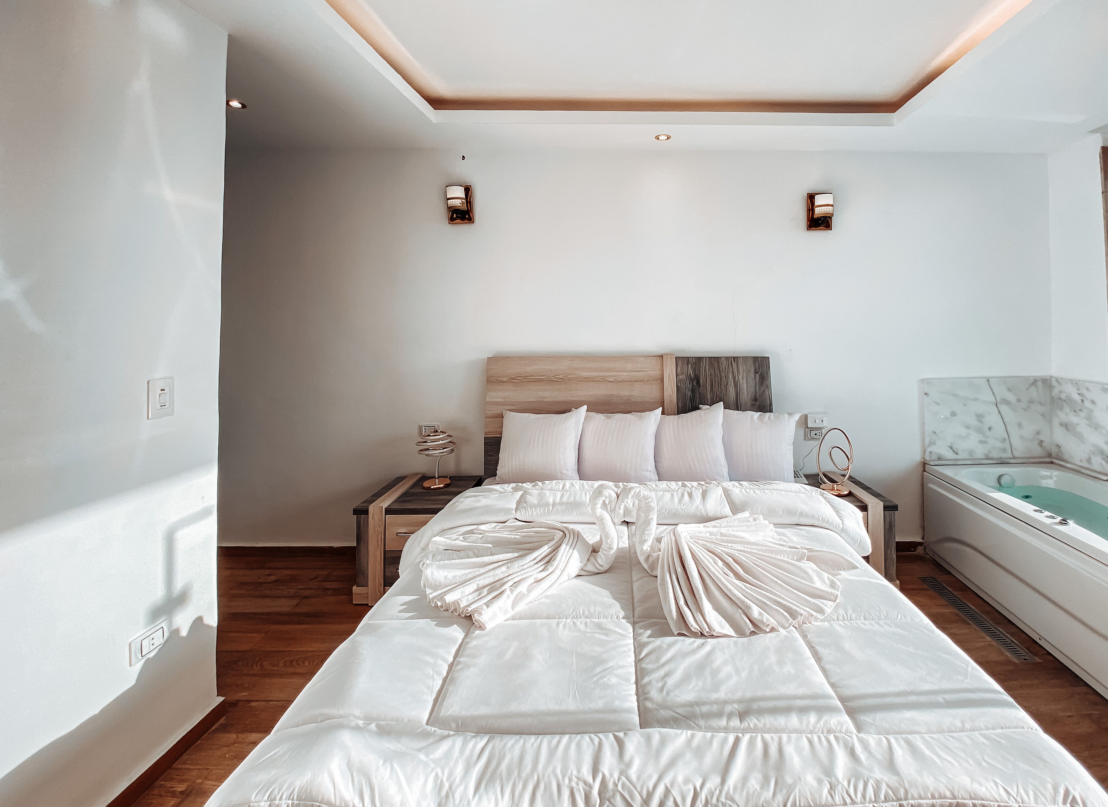 A modern hotel room with a large bed, a wooden headboard, two bedside tables with lamps, and a marble bathroom.