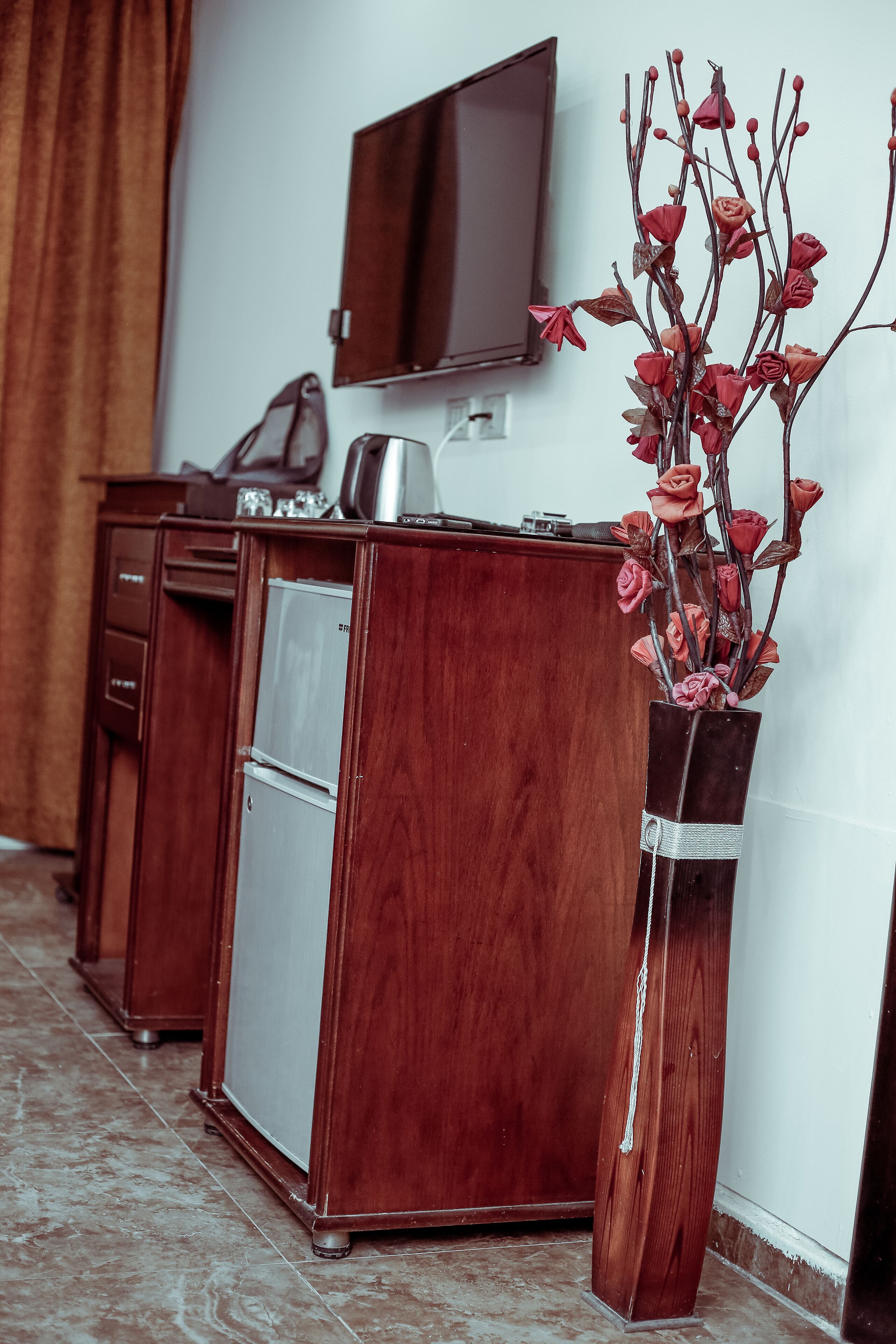 Un escritorio de madera con un televisor montado en la pared, un jarrón con flores secas y un refrigerador.
