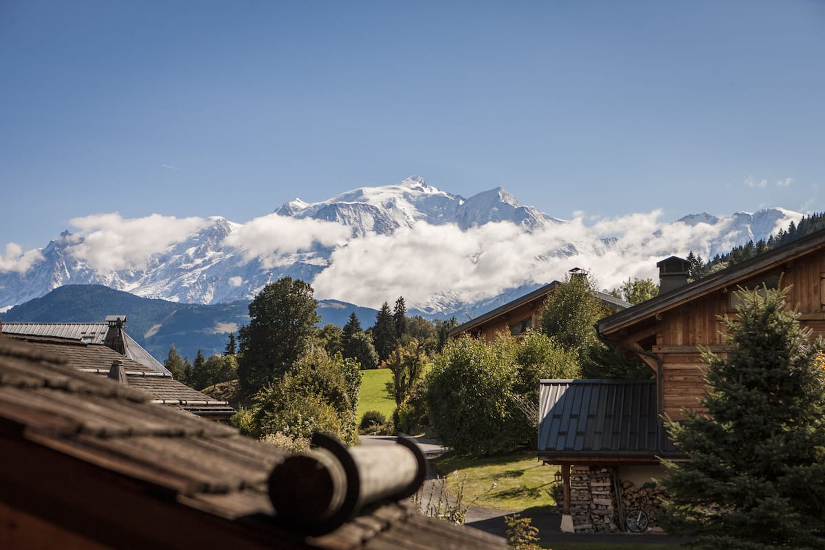 Una cabaña de madera con cimientos de piedra, rodeada de exuberante vegetación y vistas a montañas nevadas.