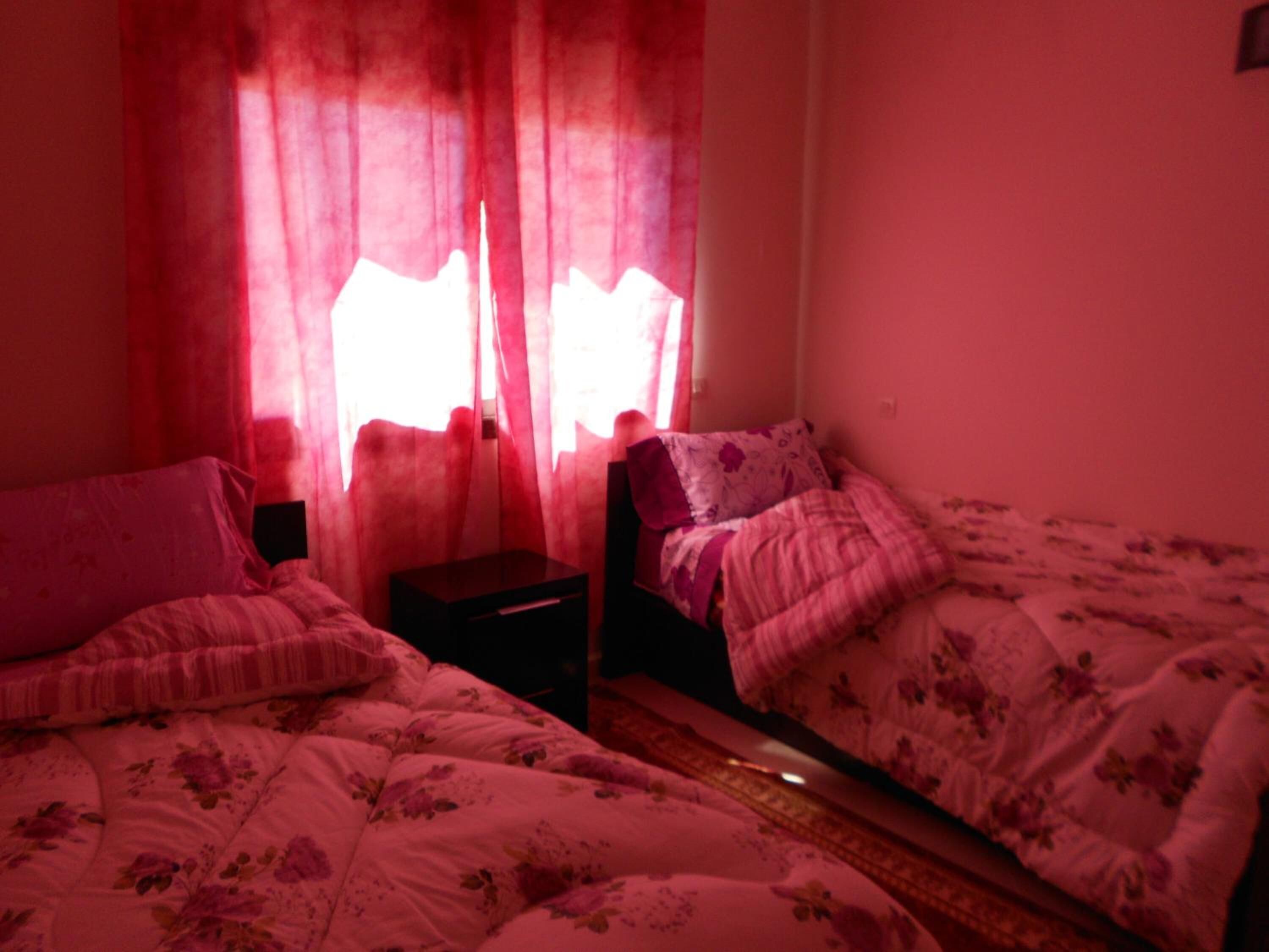 Ein Zimmer mit zwei Betten, jeweils mit floralem Bettbezug, einem Nachttisch und einem Fenster mit roten Vorhängen.