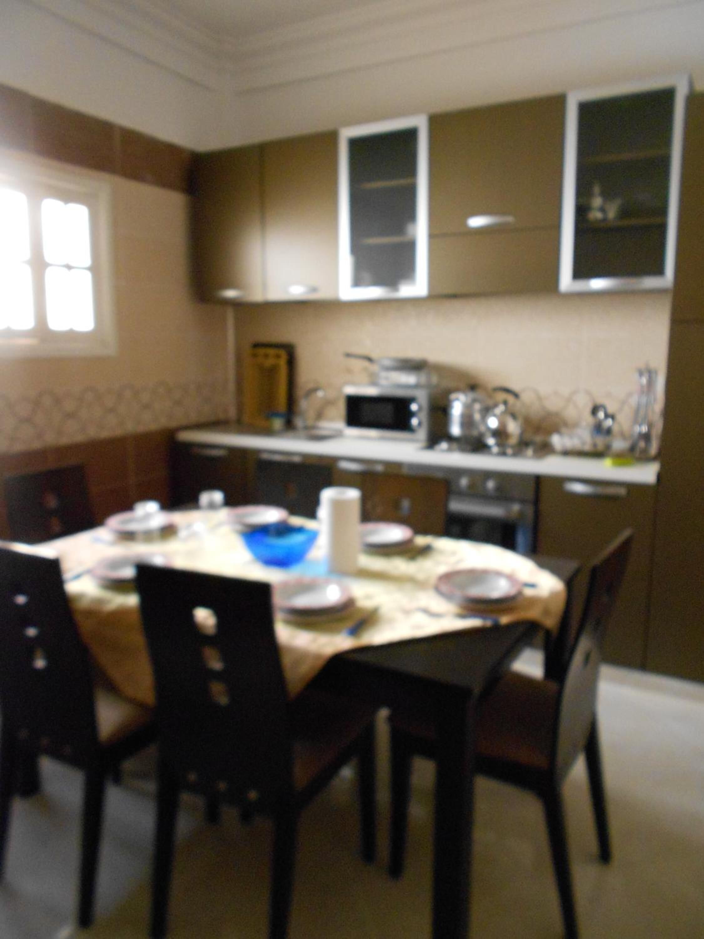 Un comedor con una mesa dispuesta para cuatro personas, rodeada de sillas. Hay un microondas y una estufa en la zona de la cocina.