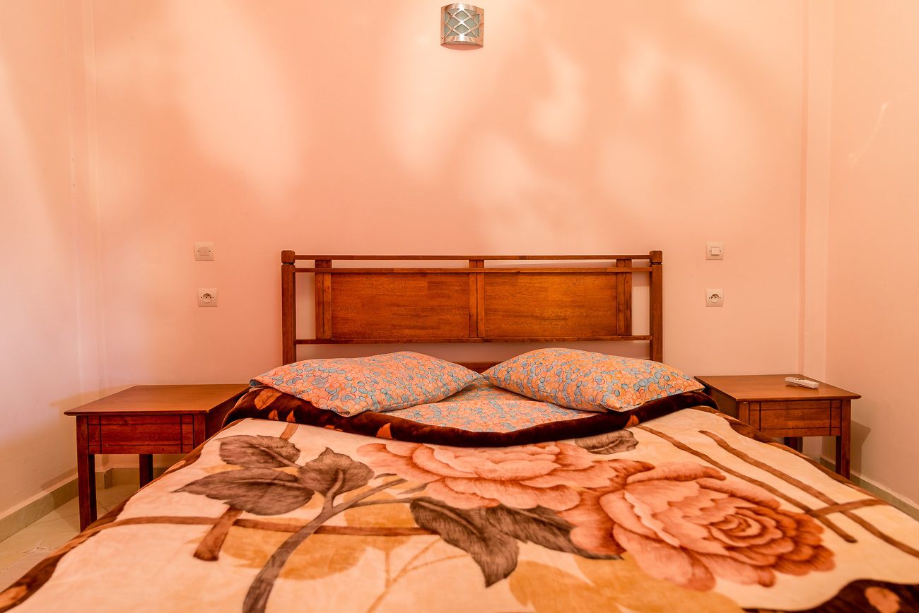 A bedroom with a wooden headboard, floral bedspread, two matching pillows, a bedside table, and a nightstand.