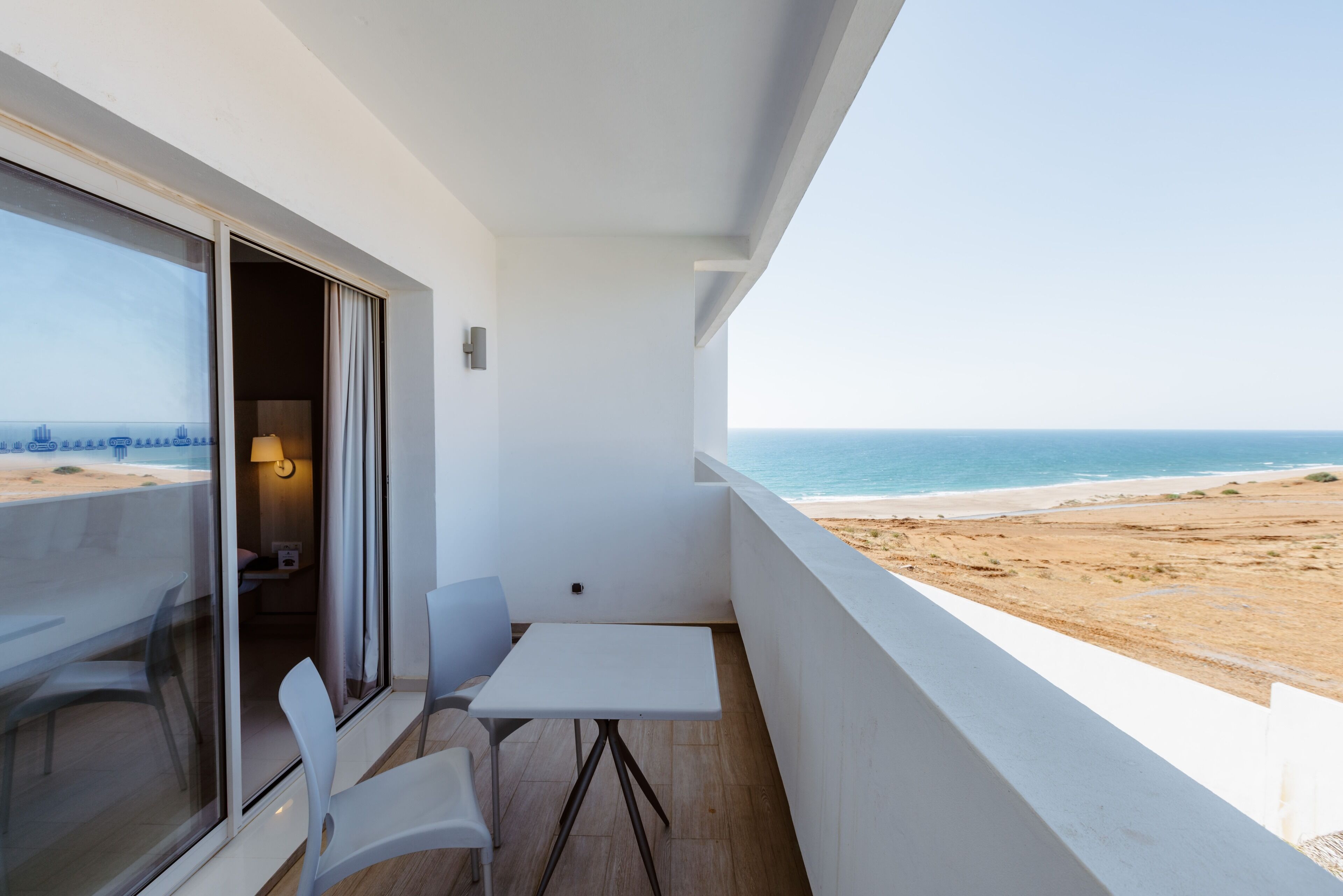 A balcony with a table and chairs, a view of the beach, and a cityscape in the background.