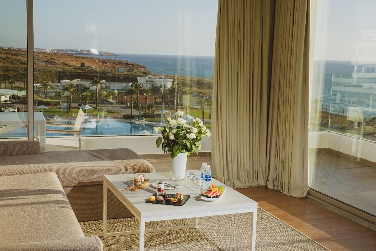 A room with a large window overlooking a pool and ocean, a white coffee table with snacks, and a beige sofa.