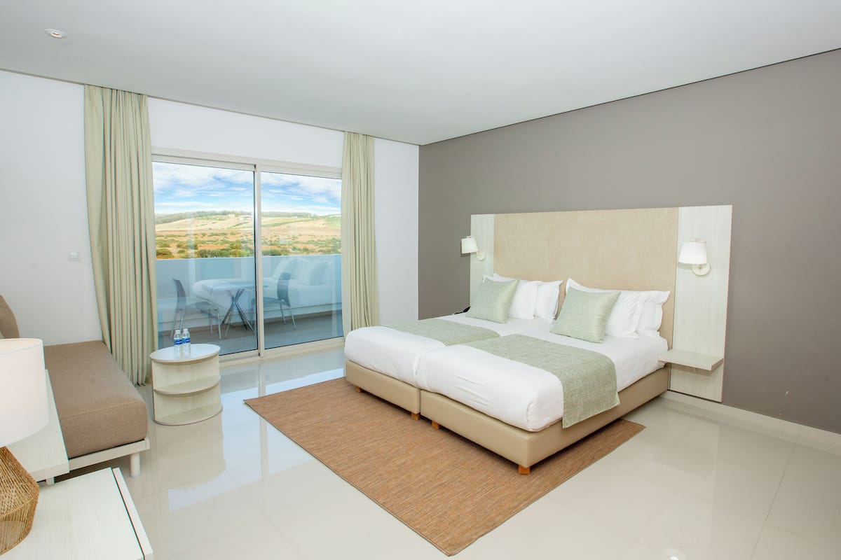A modern hotel room with a large bed, a small side table, a sofa, and a balcony with a glass door.