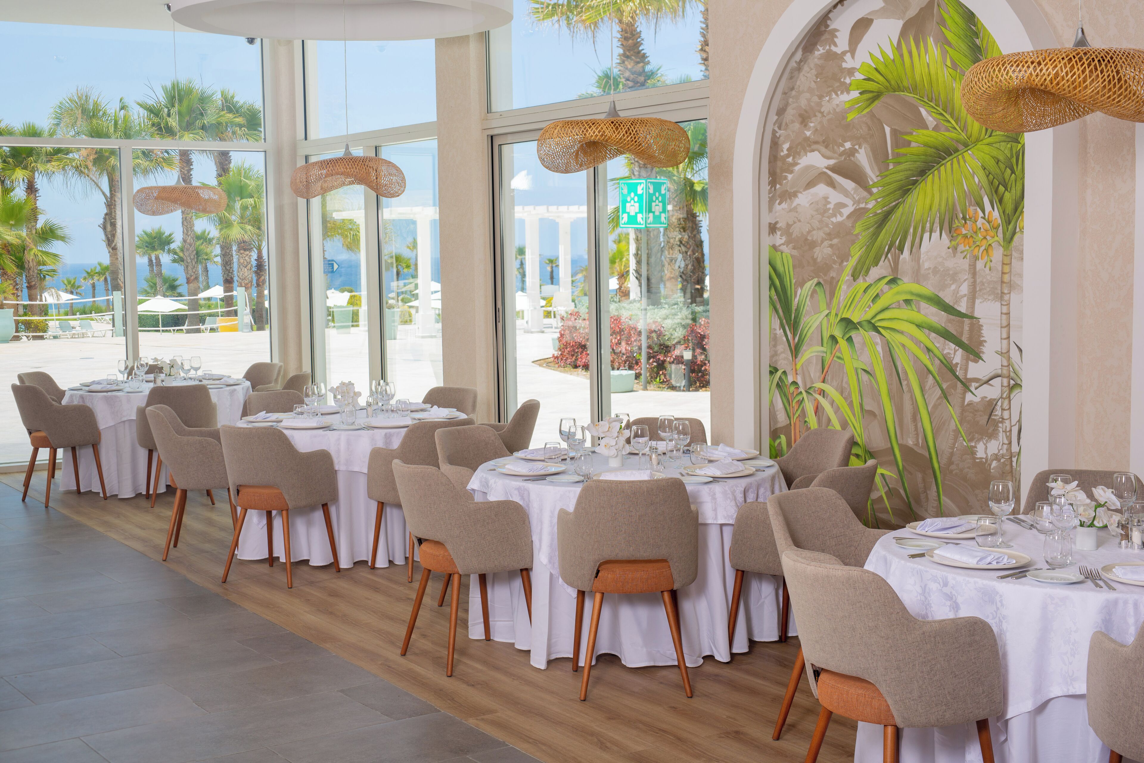 Ein Restaurant mit runden Tischen, großen Fenstern mit Blick auf Palmen und das Meer sowie einem tropischen Wandgemälde.
