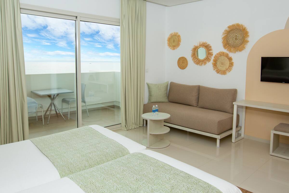 A modern hotel room with a large bed, a sofa, a small round table, and a TV mounted on the wall.