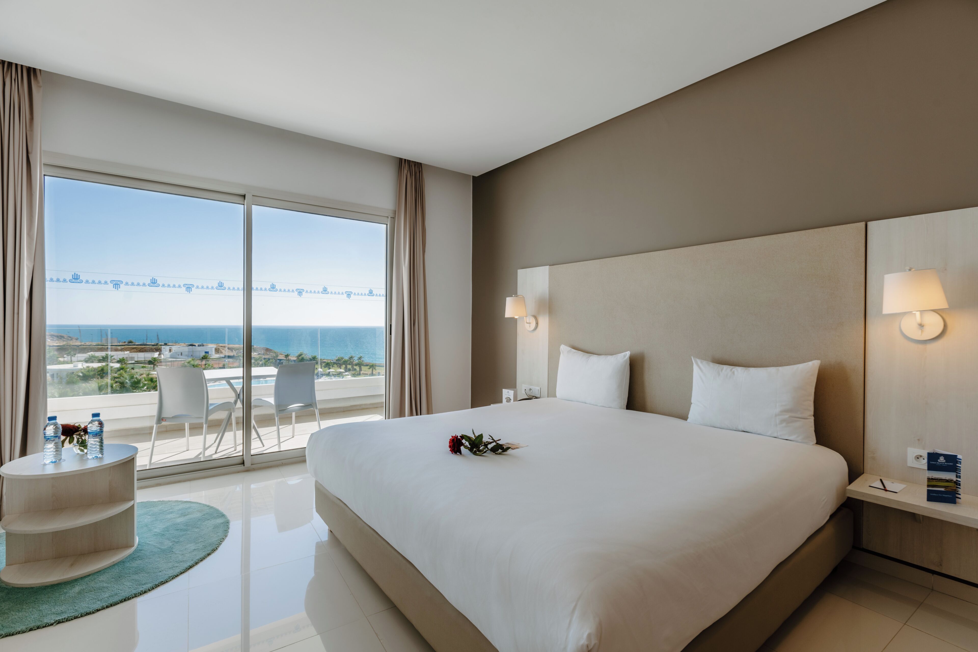 A hotel room with a large bed, a bedside table, a nightstand, a small round table with water bottles, a chair, and a view of the sea through large windows.