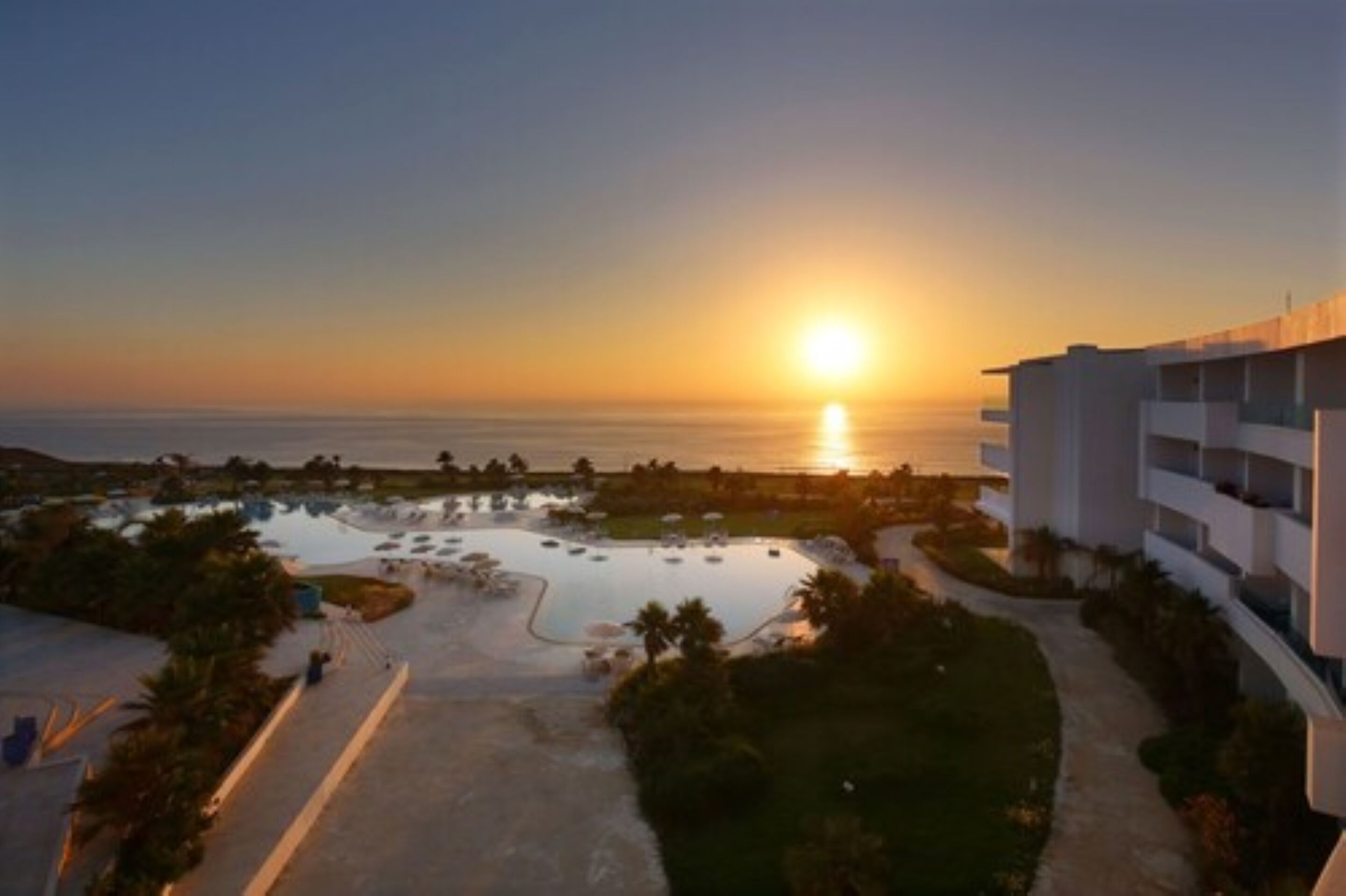 A resort with a swimming pool, a building with multiple balconies, and a view of the sunset over the ocean.