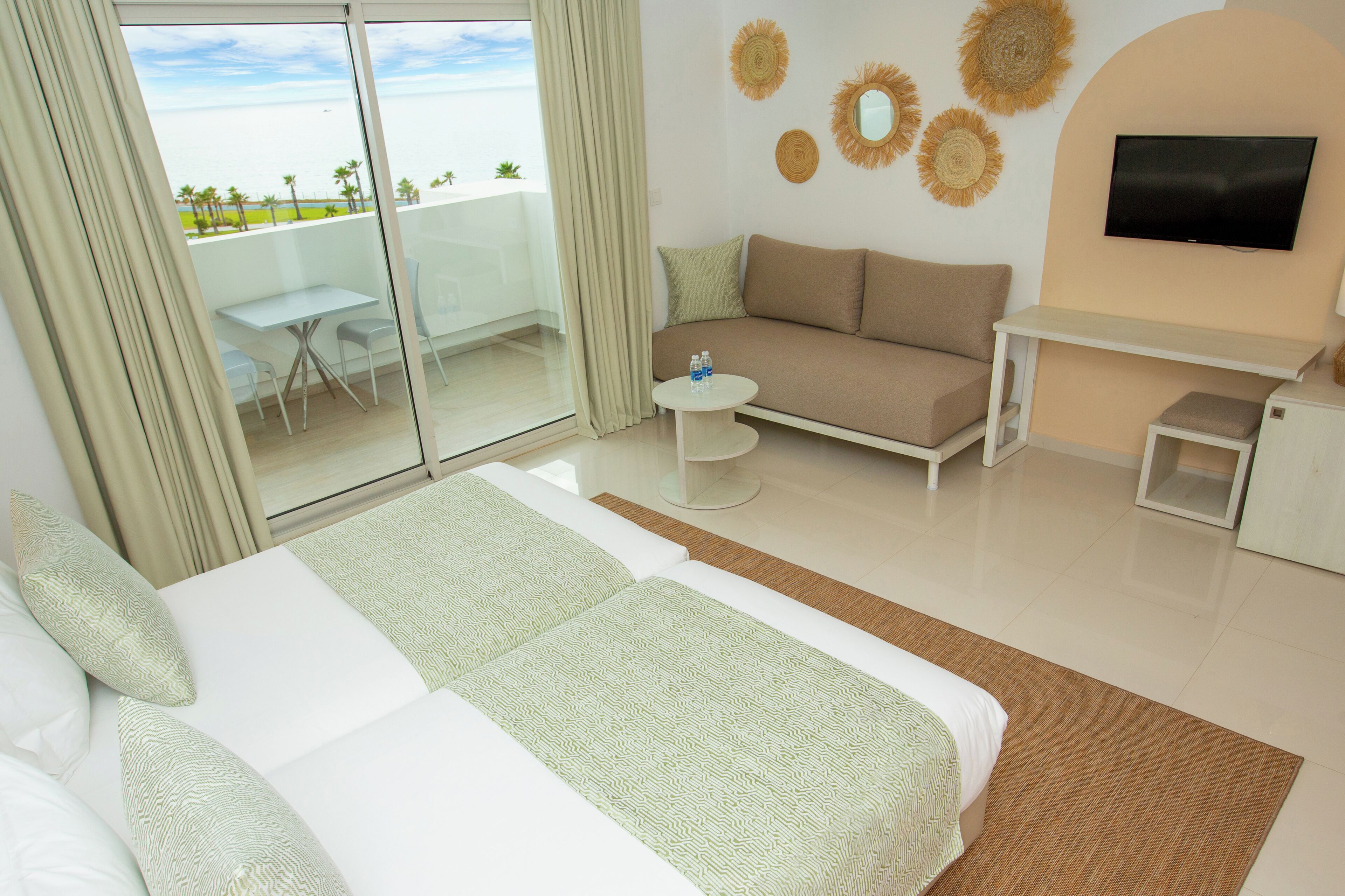 A hotel room with a bed, a sofa, a TV, a small table, and a balcony with a view of the ocean.
