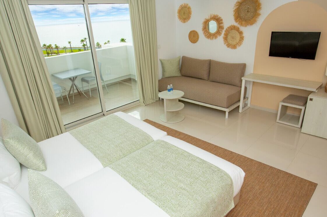 A hotel room with a large bed, a sofa, a small table, and a TV. There is a balcony with a glass door and a view of the beach.