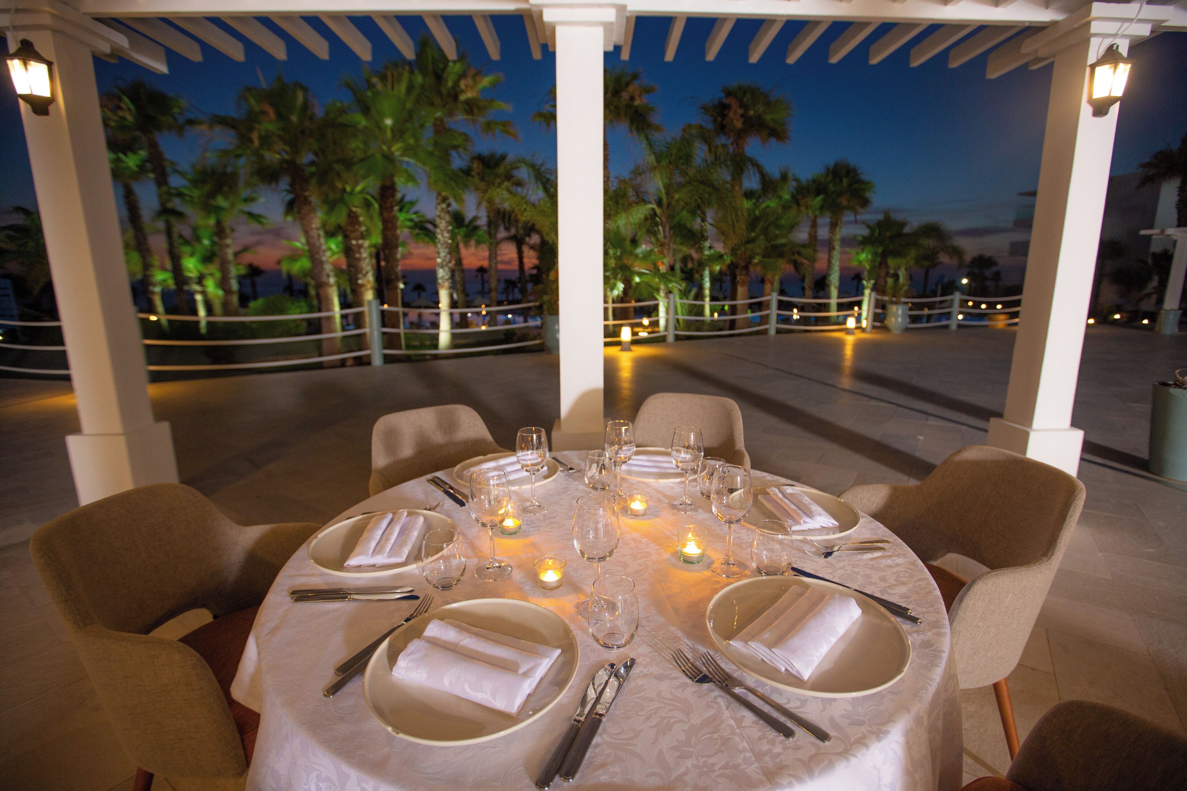 Una mesa dispuesta para comer, con velas, palmeras y vista al océano de fondo.