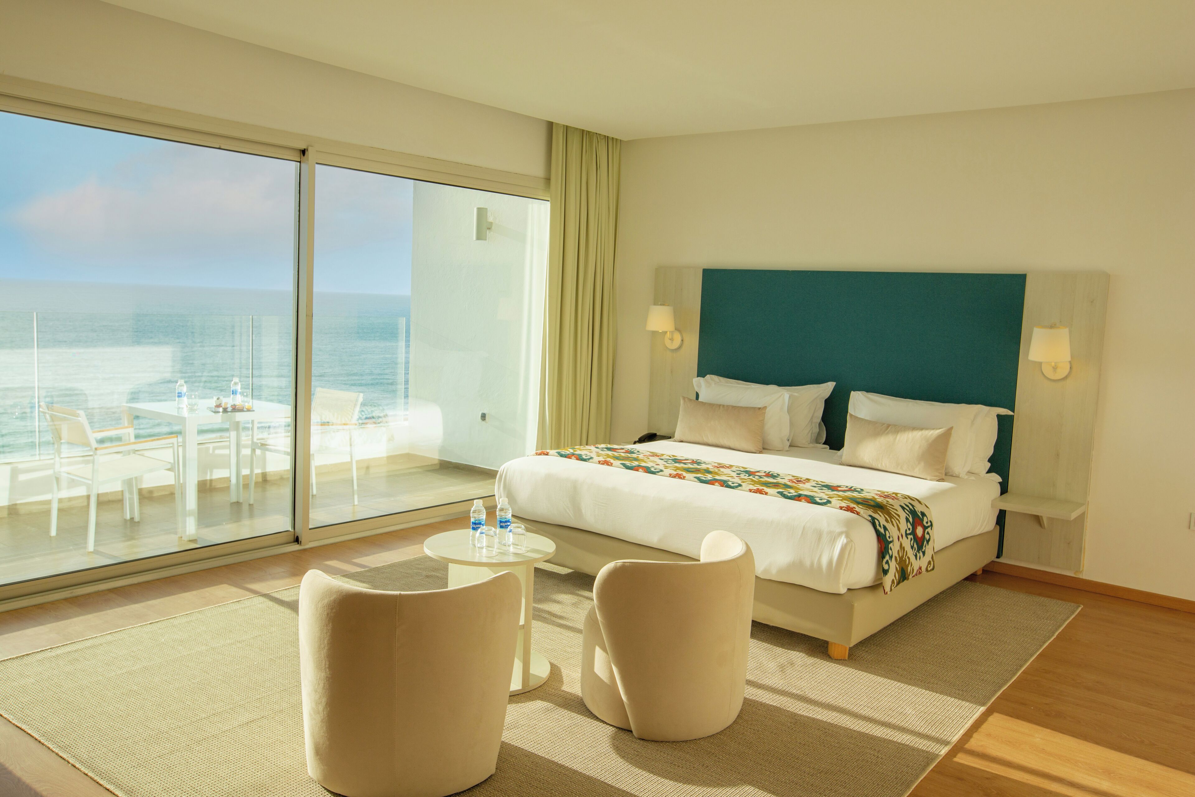 Een hotelkamer met een groot bed, een balkon met uitzicht op zee, een klein rond tafeltje en twee stoelen.