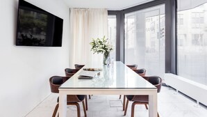 A modern meeting room with a glass table, wooden chairs, a vase with flowers, a mounted TV, and large windows with blinds. - Skyreach Bizzstay - Wohnen Am Main (Frankfurt)
