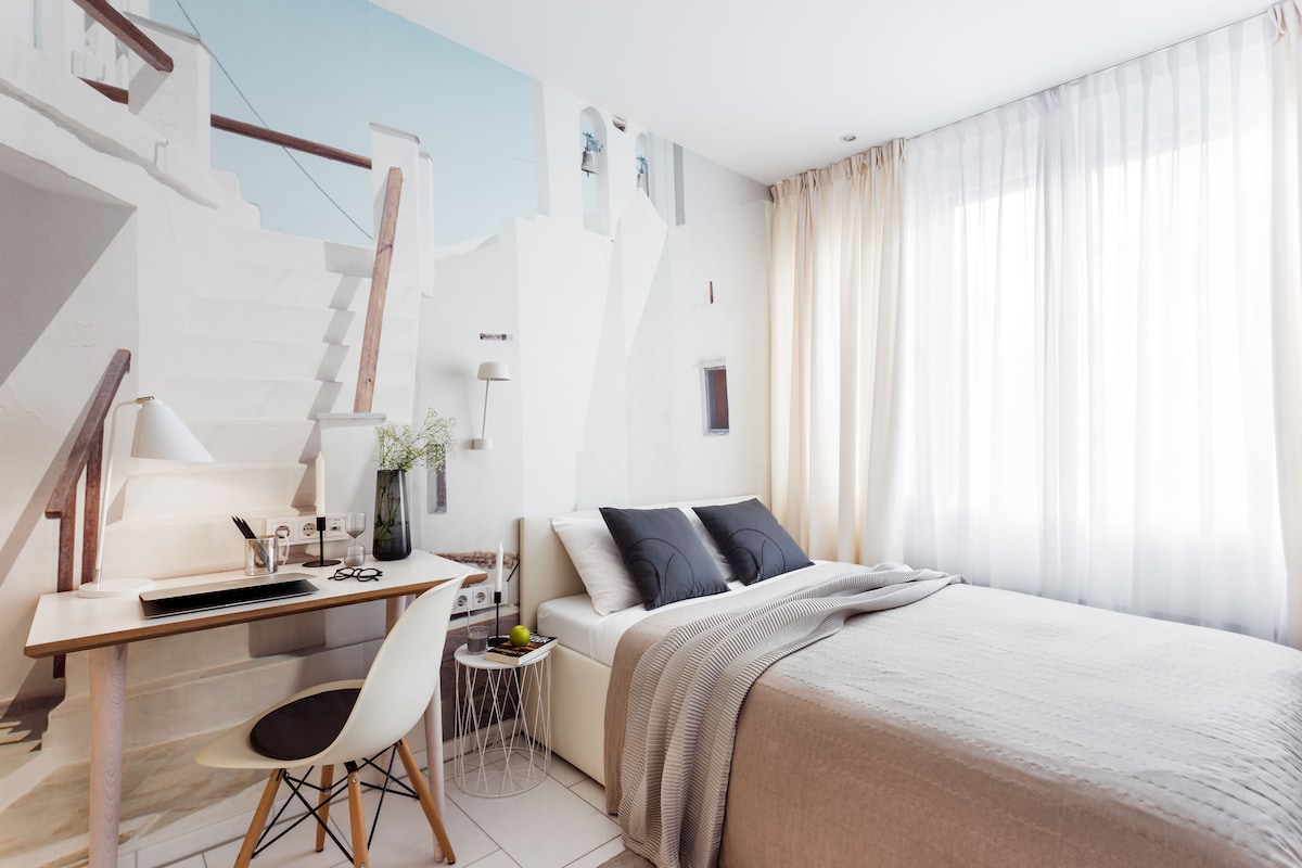 A modern hotel room with a bed, desk, chair, and a small side table.