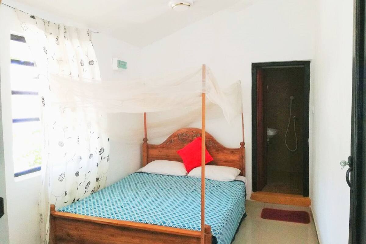 A single bed with a wooden frame and a blue patterned bedspread. There is a white canopy over the bed, and a red pillow is visible. A door with a shower is to the right of the bed.