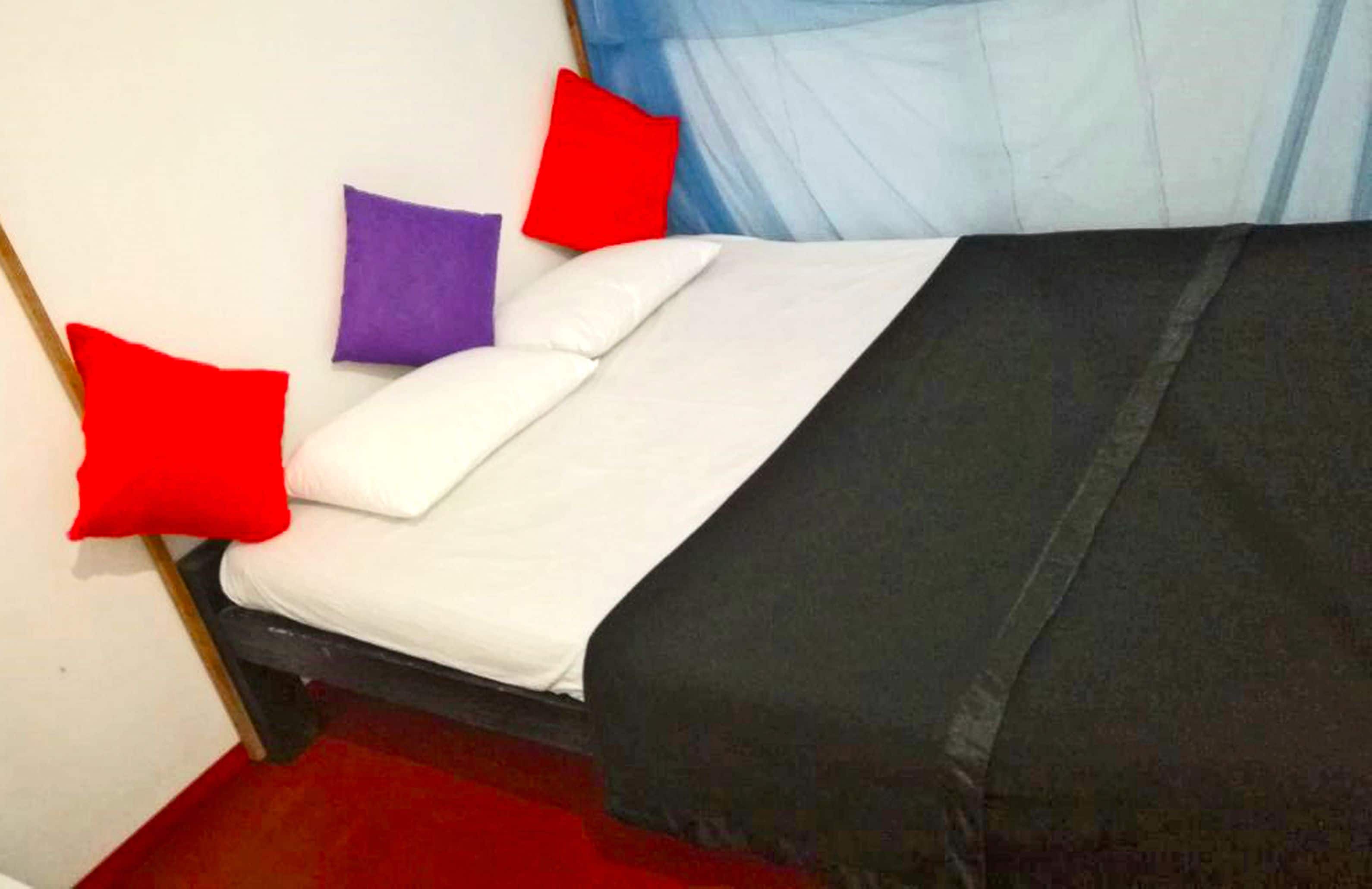 A bed with white and black bedding, red and purple pillows, and a blue sheer curtain.