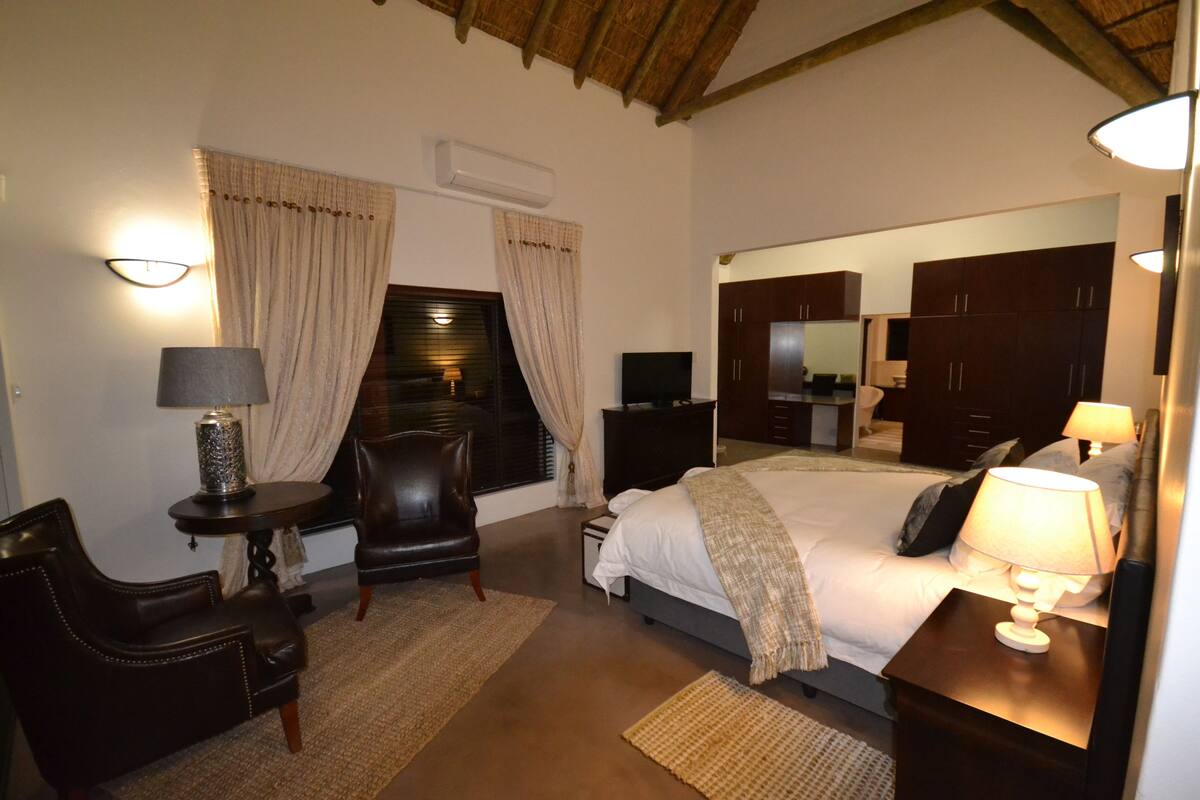 A bedroom with a large bed, a desk with a lamp, a chair, a TV, and a wardrobe.