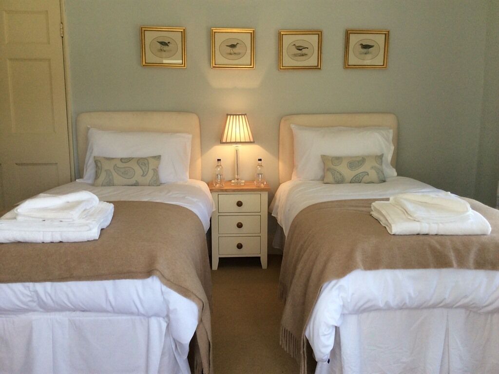 A hotel room with two beds, each with a pillow and a folded towel. There is a nightstand with a lamp and two bottles. The walls have framed pictures of birds.