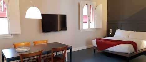 A room with a bed, a dining table with chairs, a TV, and a window with curtains.