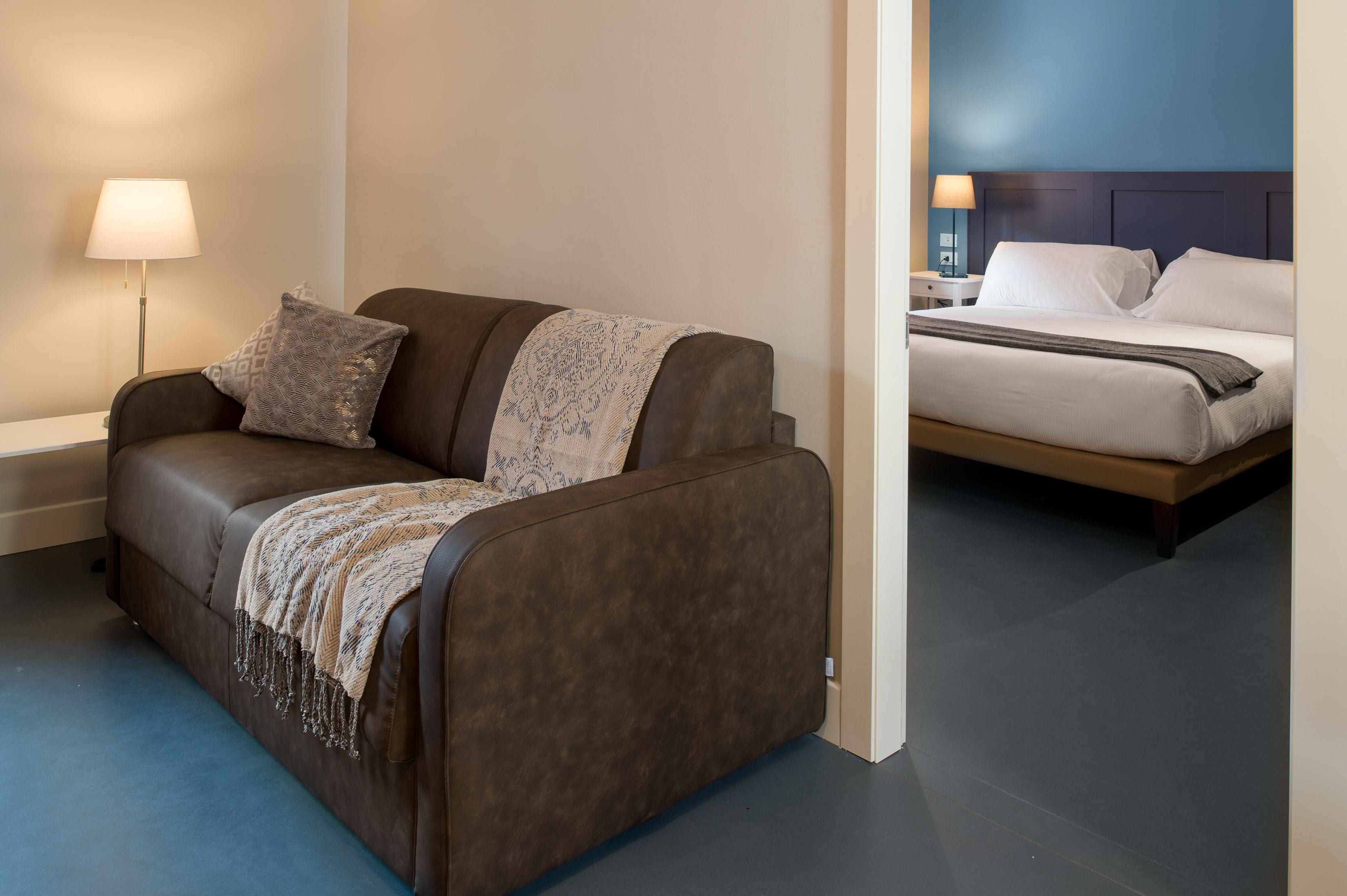 A modern hotel room with a brown sofa, a bedside table with a lamp, and a bed with white linens.
