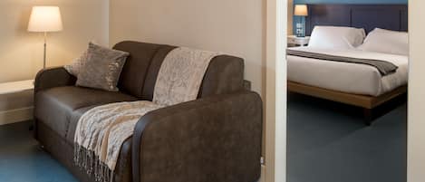 A modern hotel room with a brown sofa, a bedside table with a lamp, and a bed with white linens.