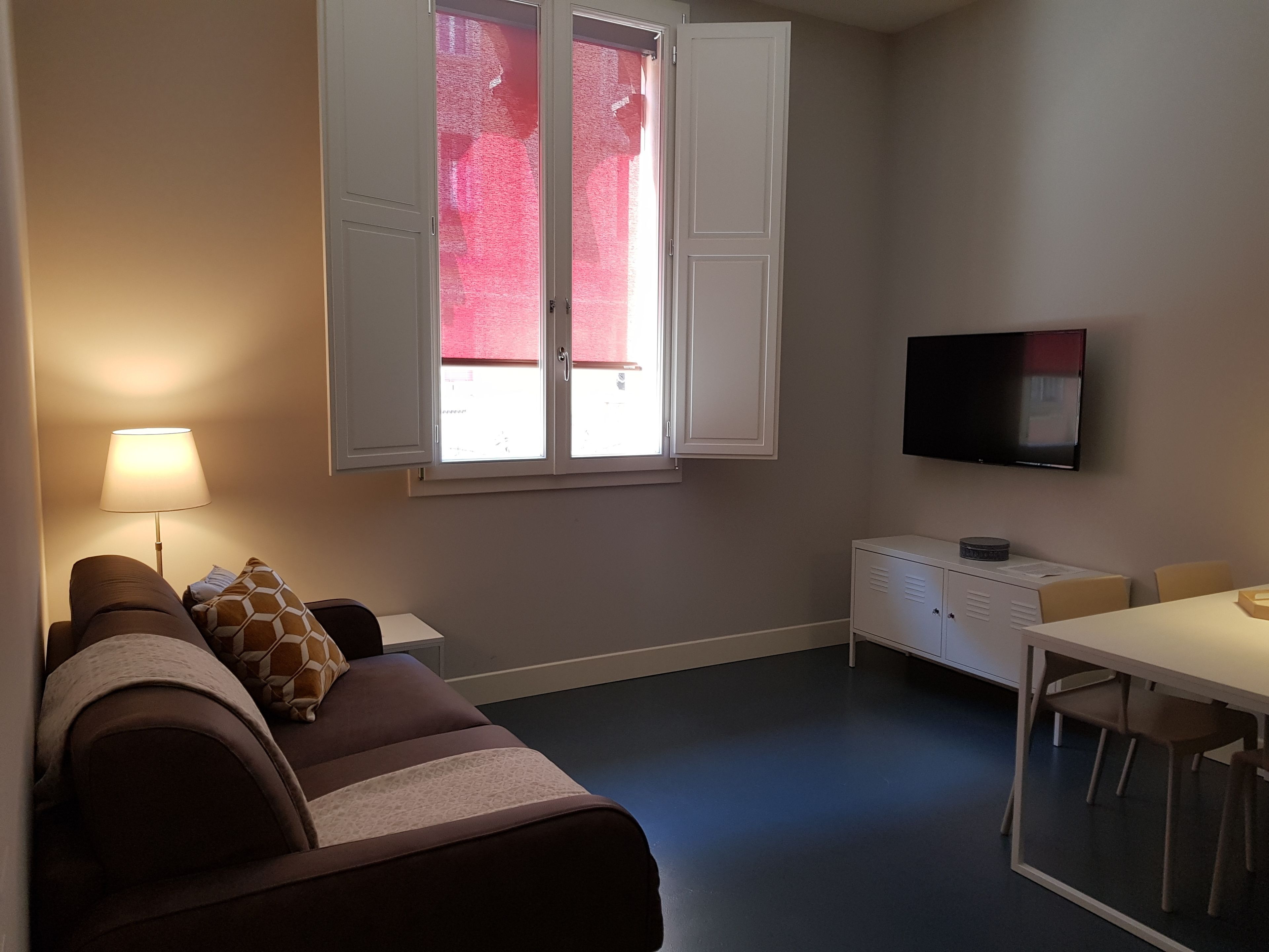 Una habitación con sofá, televisión montada en la pared, ventana con contraventanas blancas y una pequeña mesa con sillas.