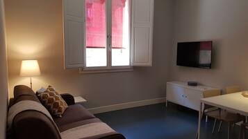 A room with a sofa, a TV mounted on the wall, a window with white shutters, and a small table with chairs.