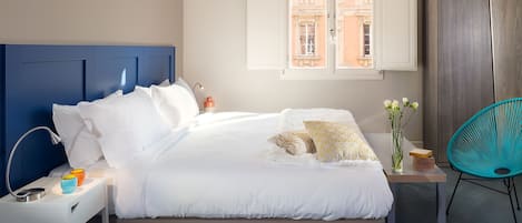 Een keurig opgemaakt bed met witte lakens en een blauw hoofdeinde, een nachtkastje met een lamp en een kleine vaas, en een uitzicht op een gebouw door een raam.