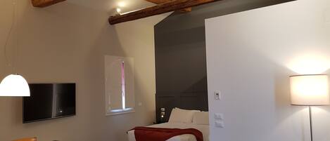 Una habitación moderna con vigas de madera a la vista, una cama con manta roja, un sofá de cuero negro, una mesa de comedor con sillas y un televisor de pantalla plana montado en la pared.