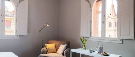 A bedroom with a bed, a grey armchair with yellow pillows, a desk with a lamp, a vase with sticks, and a teapot.