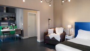 A modern hotel room with a bed, bedside table, lamp, and a sofa. There is a kitchenette with a stove, sink, and cabinets. - Almarossa (Bologna)