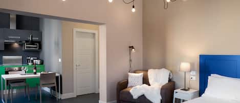A modern hotel room with a bed, bedside table, lamp, and a sofa. There is a kitchenette with a stove, sink, and cabinets.
