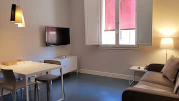 Ein modernes Wohnzimmer mit Essbereich, einem flachbildfernseher an der Wand, einem Fenster mit Rollläden und einem Sofa mit gemustertem Kissen.