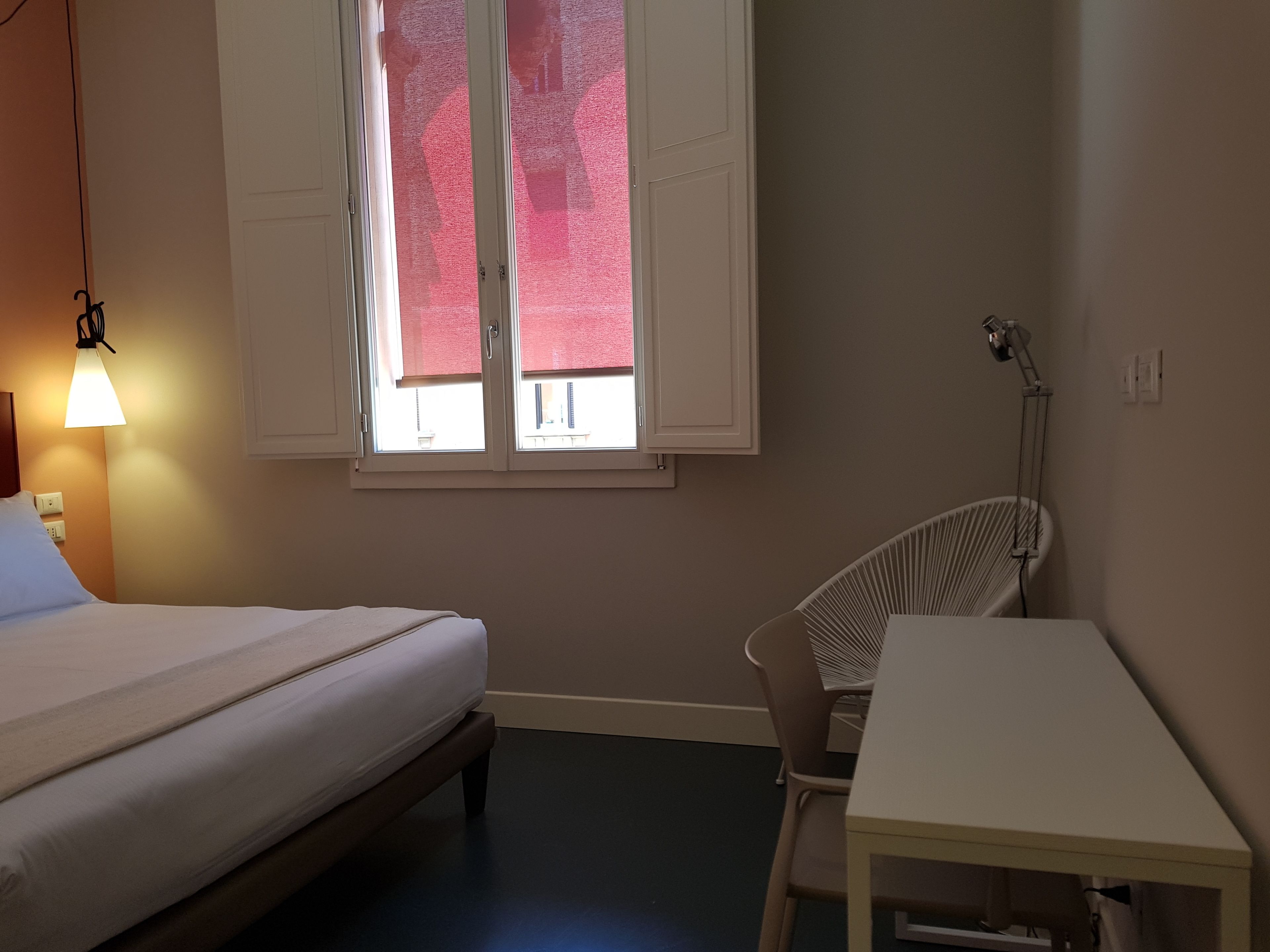A bedroom with a bed, a desk, a chair, a window with blinds, and a lamp.